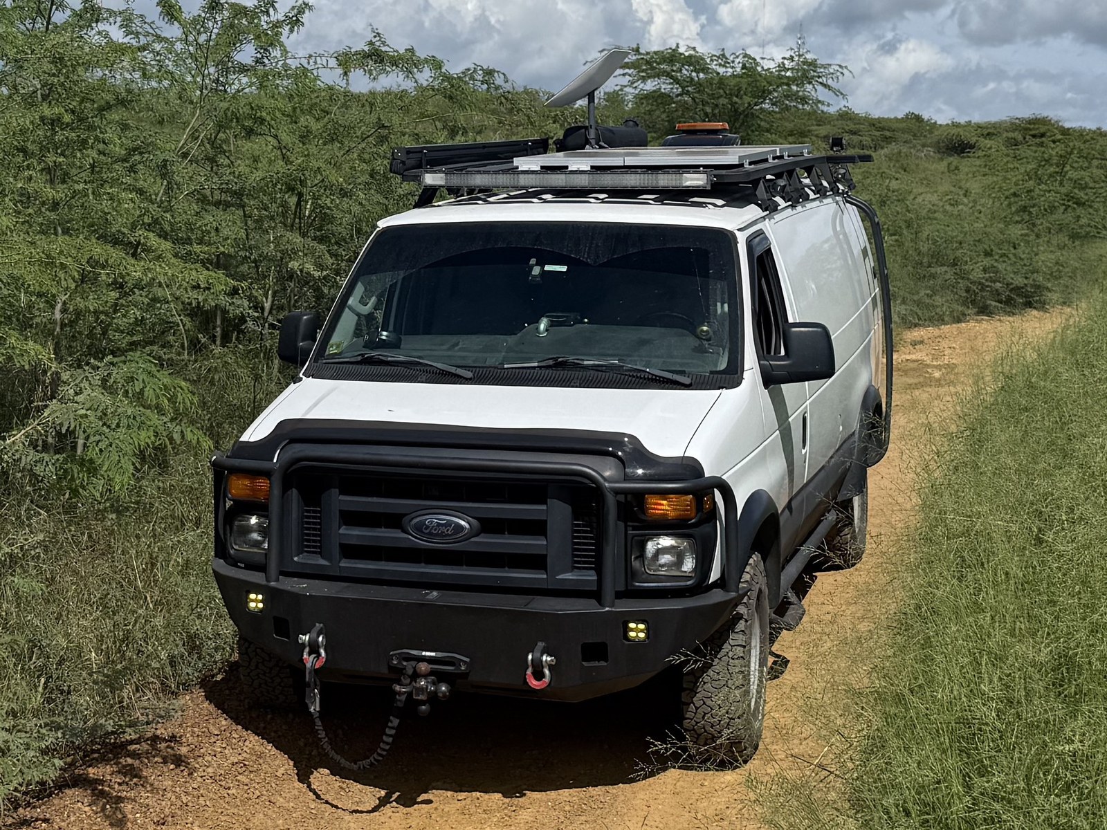 The white Ford E-Series van on a narrow dirt road, custom front bumper and roof rack, Starlink dish mounted on the rack
